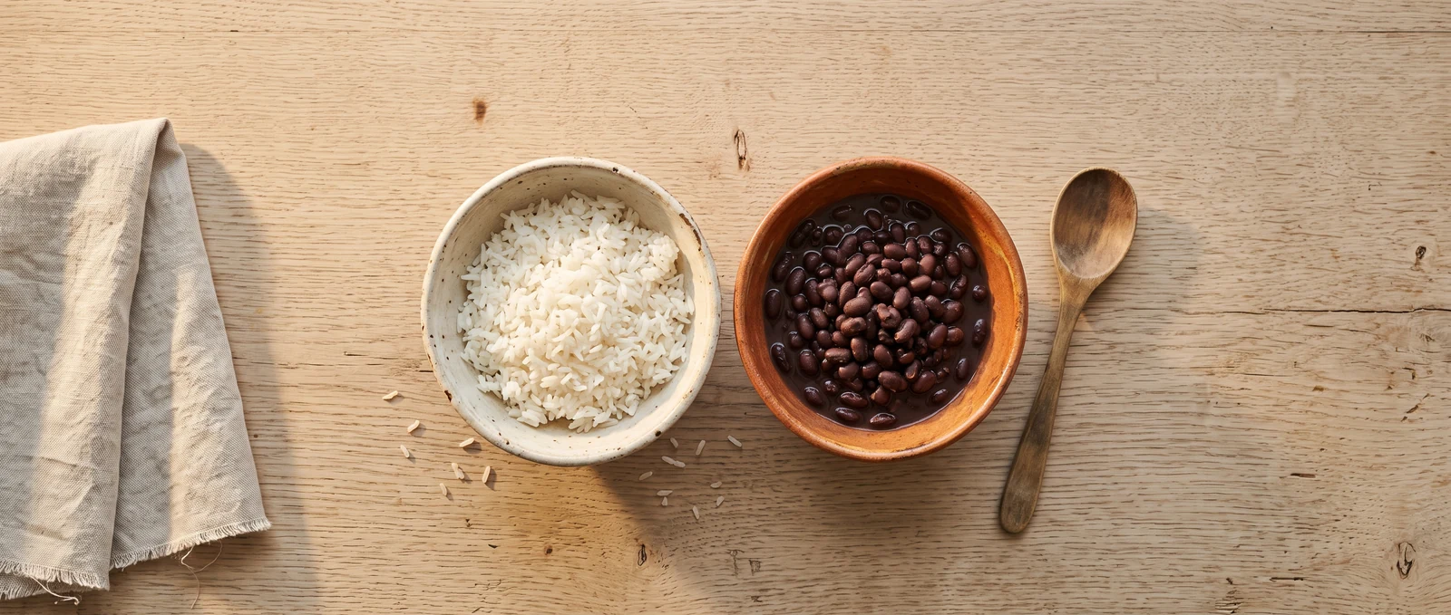 A bowl of white rice and a bowl of black beans side by side on a pale oak table, seen from above.