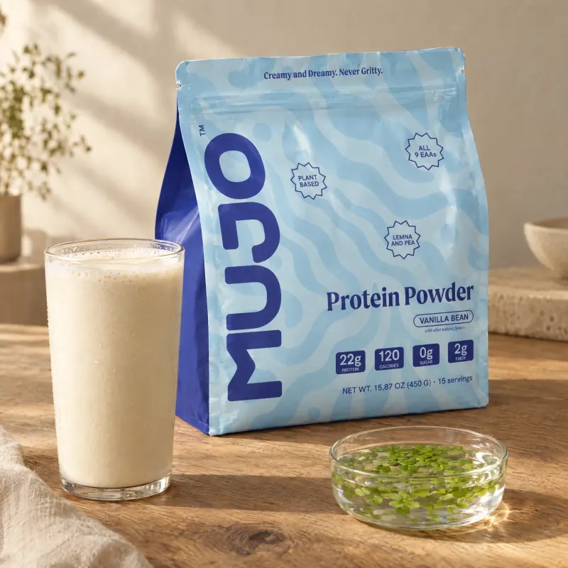 A pouch of Mujo Protein Powder beside a glass of vanilla shake and a small dish of Lemna leaves.